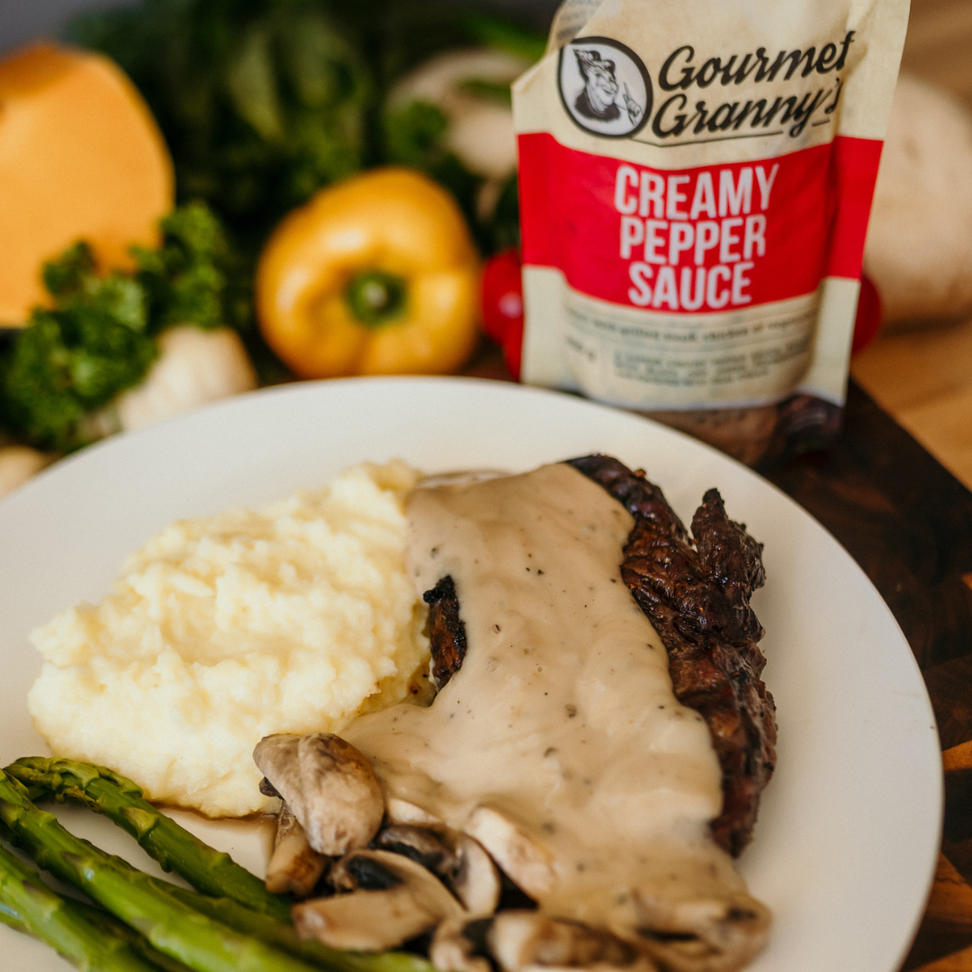 Plate of food with creamy pepper sauce, asparagus, and mashed potatoes, with a packet of Gourmet Granny Creamy Pepper Sauce in the background.
