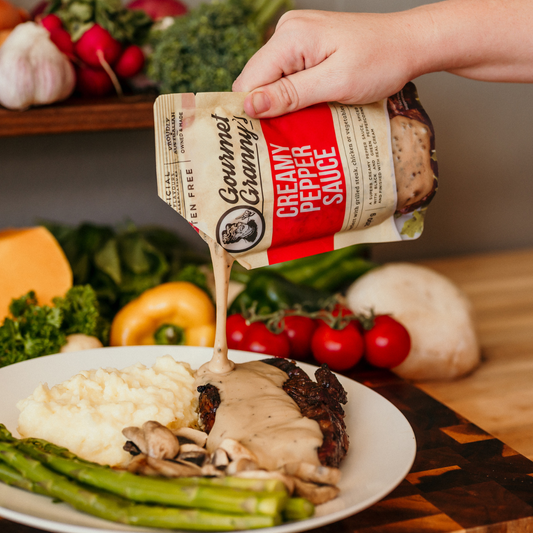 Hand pouring Gourmet Granny's creamy pepper sauce over a plate of food with vegetables in the background.