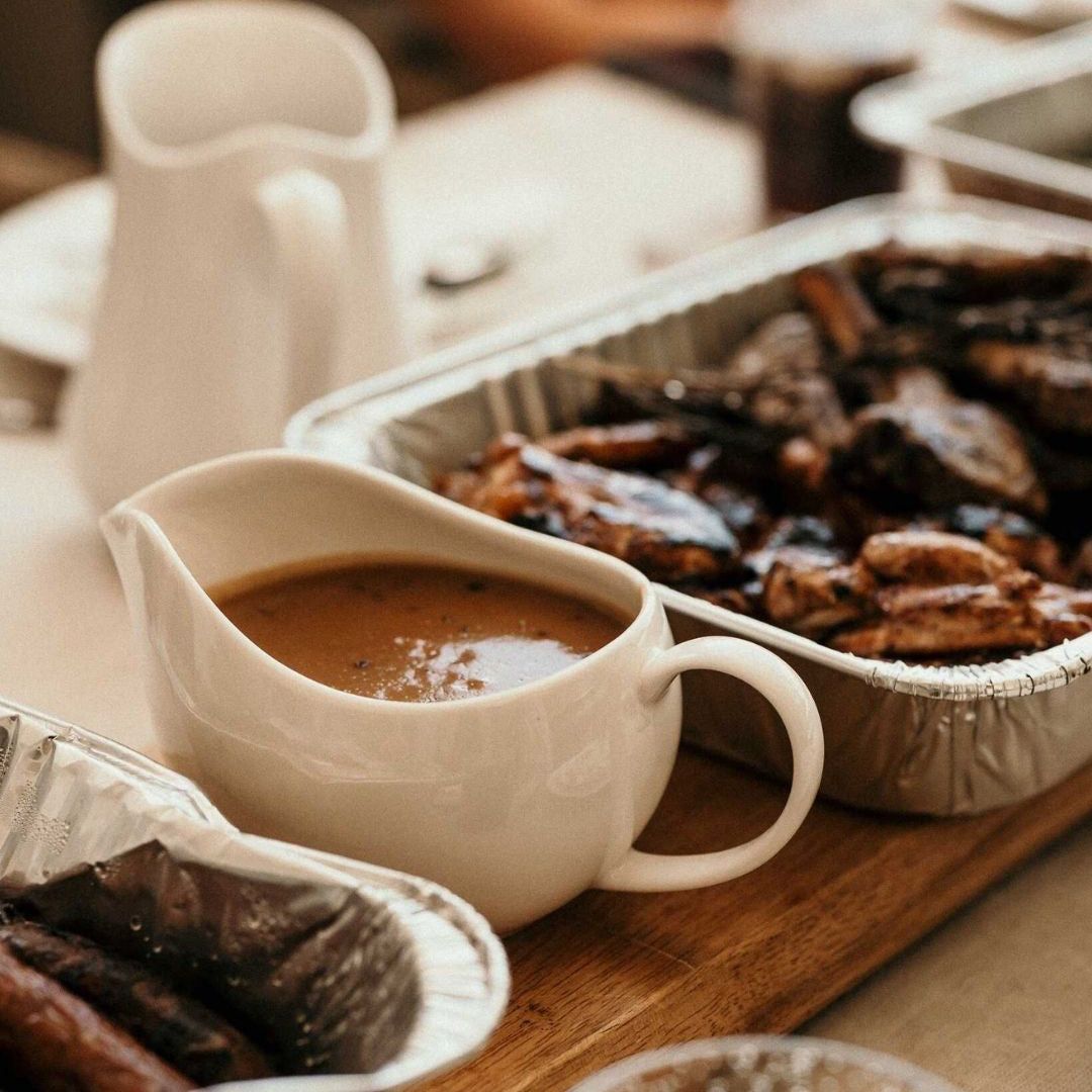 White ceramic gravy boat filled with  gravy, on a wooden surface with roast meats.