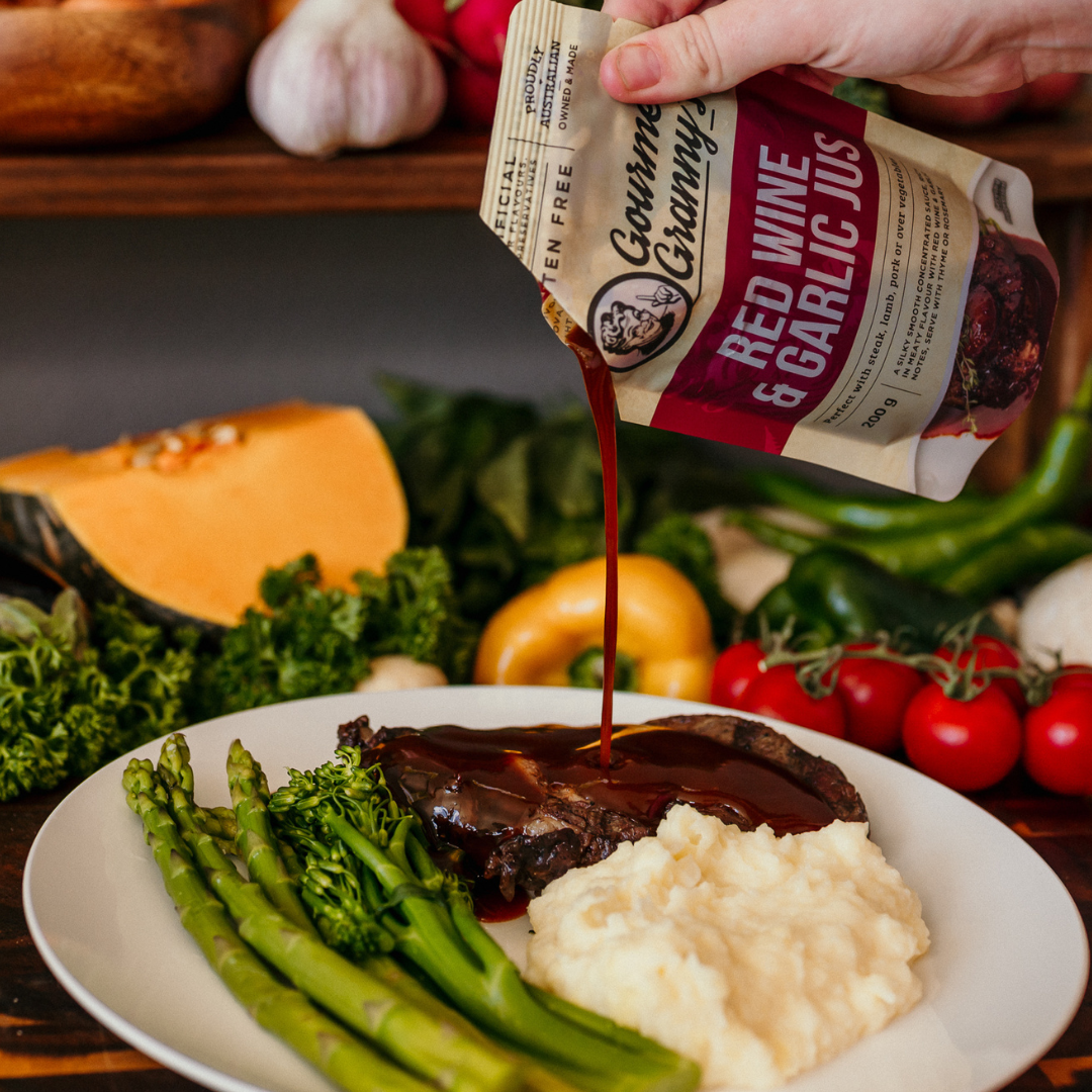 Person pouring red wine & garlic jus over a plate of food with vegetables in the background