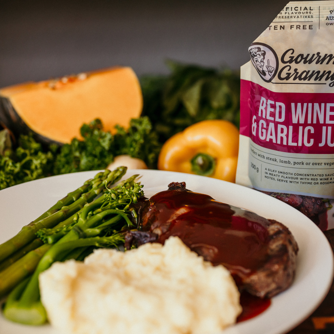Plate of food with steak, asparagus, and mashed potatoes, accompanied by a packet of red wine and garlic jus.