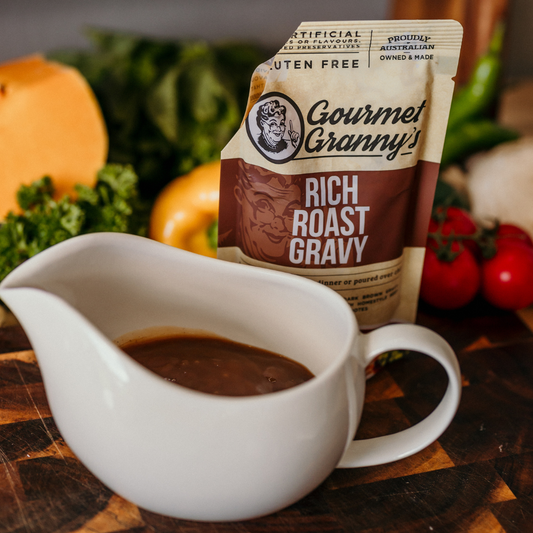 Gourmet Granny's Rich Roast Gravy packet with a white gravy boat filled with gravy on a wooden surface with vegetables in the background.